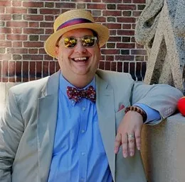 Kyle standing outside wearing a hat and sunglasses with this left arm resting on a ledge.